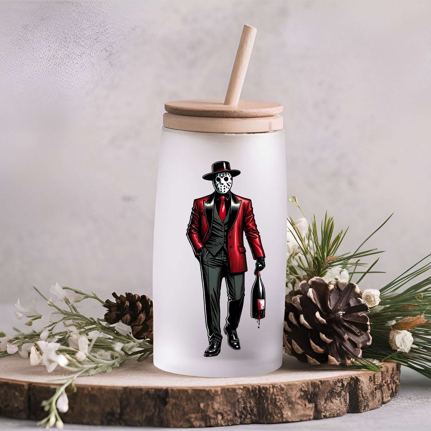 A frosted glass tumbler with a wooden lid and straw, featuring a horror character design. The character wears a red and black pinstripe suit with a red tie and a black hat, holding a dripping bottle of wine. The tumbler is placed on a rustic surface with pinecones and greenery in the background.