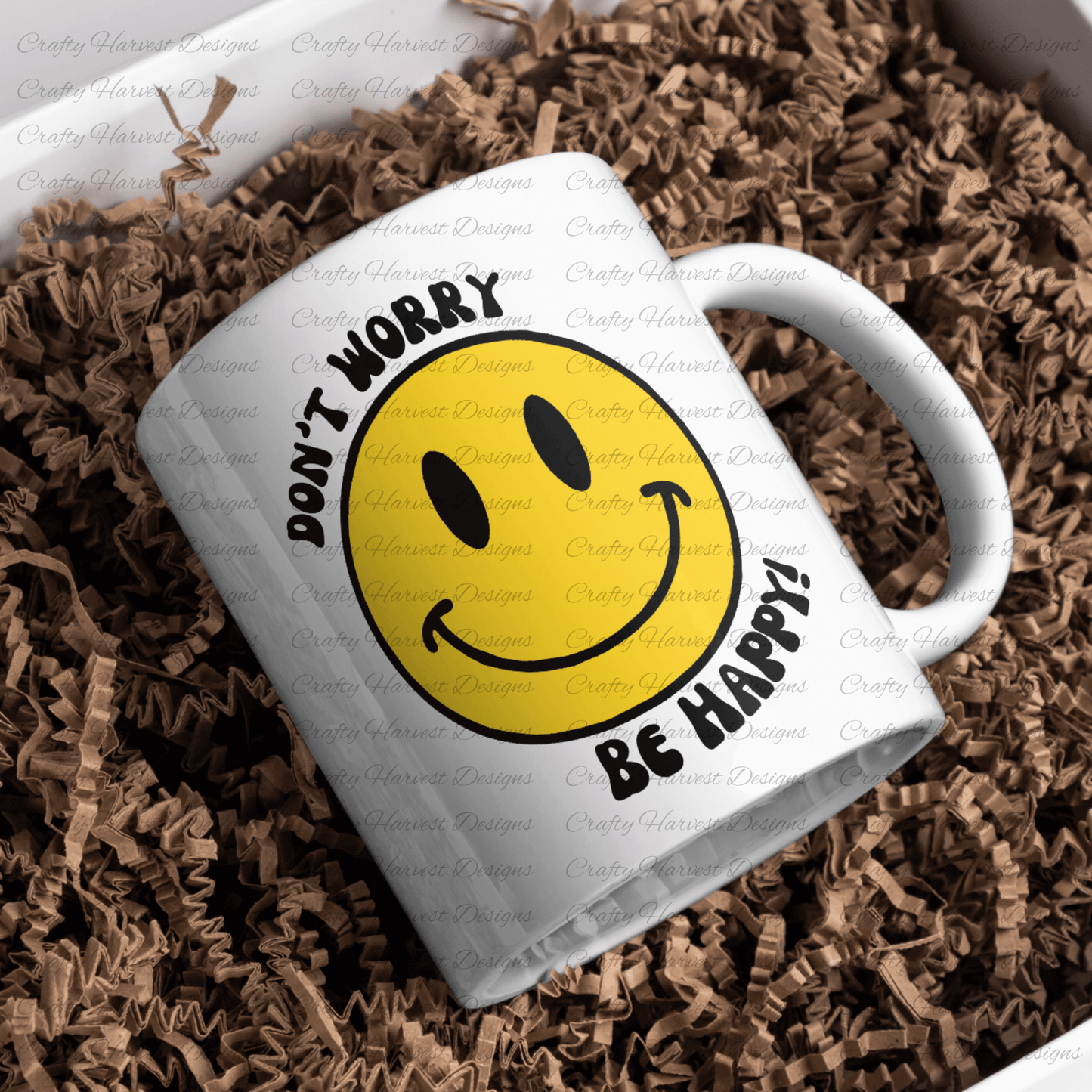 White ceramic mug featuring a classic yellow smiley face design with the phrase ‘Don’t Worry, Be Happy!’ in black text. The mug is placed on a bed of crinkled brown paper filler.