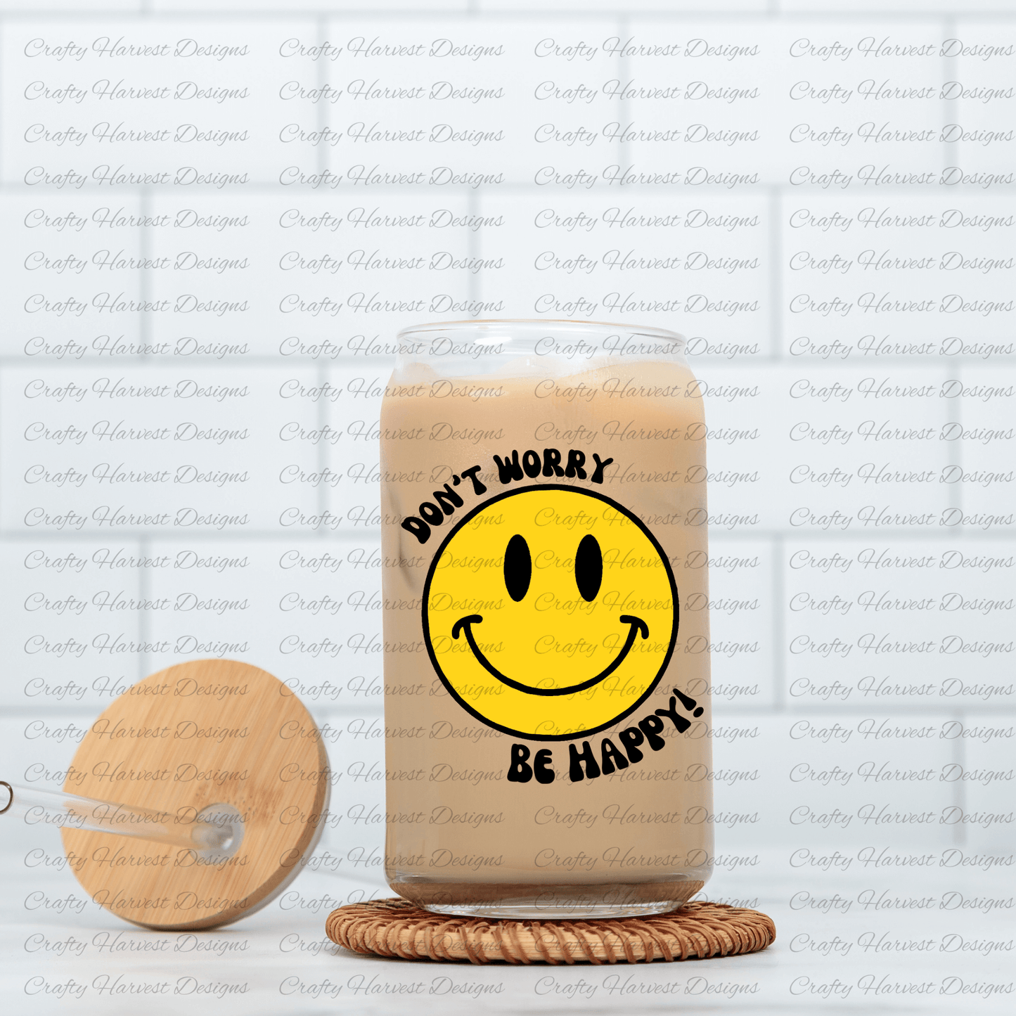 Glass can tumbler filled with iced coffee, featuring a classic yellow smiley face design with the phrase ‘Don’t Worry, Be Happy!’ in black text. The tumbler is placed on a woven coaster, with a wooden lid and glass straw resting beside it.