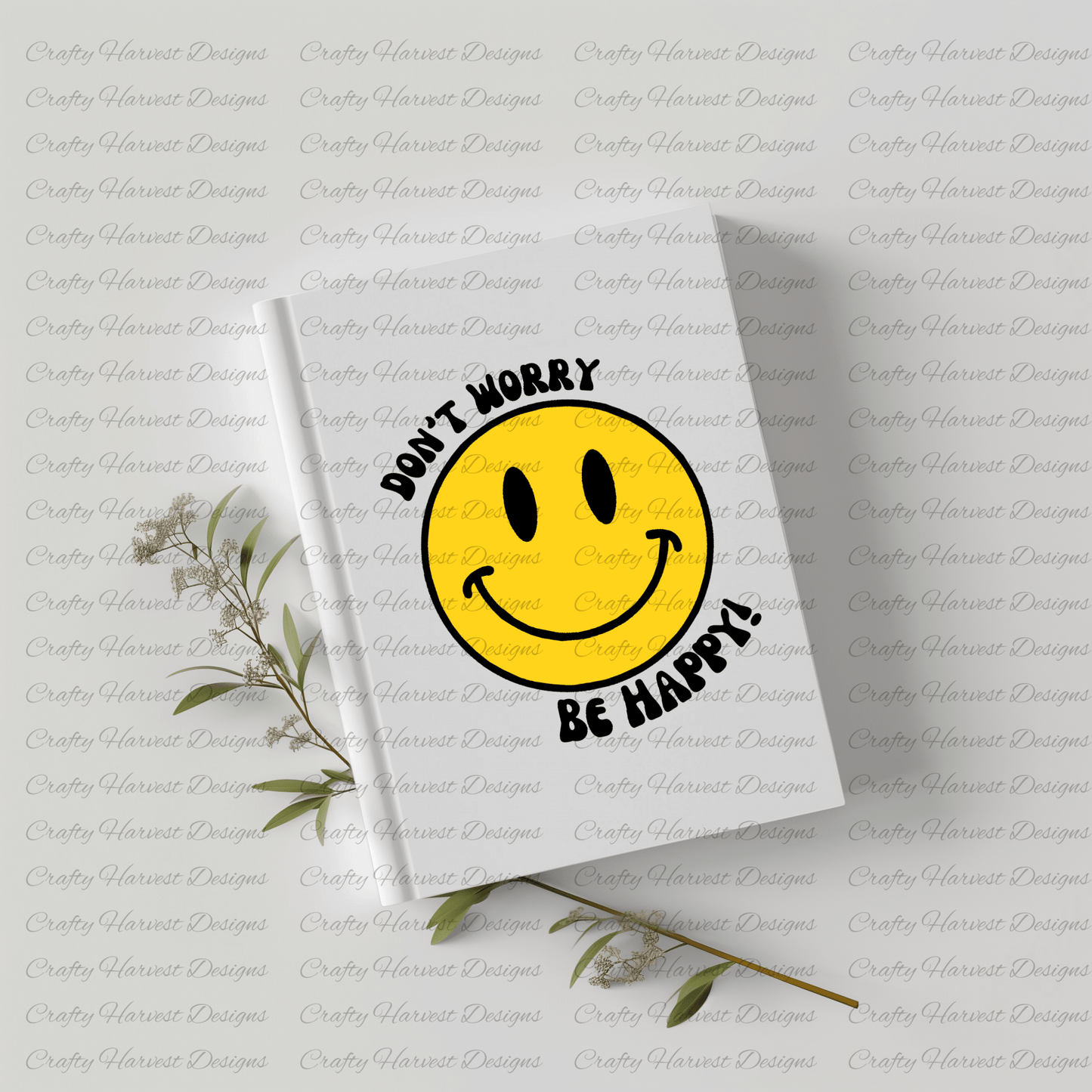 White hardcover notebook featuring a classic yellow smiley face design with the phrase ‘Don’t Worry, Be Happy!’ in black text. The notebook is placed on a light-colored surface with dried plant stems beside it.