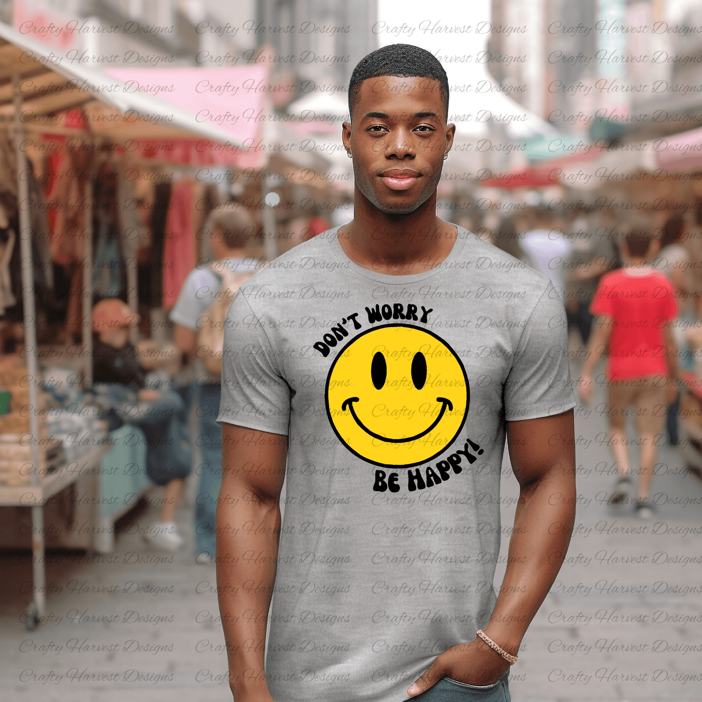 Man walking through an outdoor market wearing a gray t-shirt featuring a classic yellow smiley face design with the phrase ‘Don’t Worry, Be Happy!’ in black text. The background is slightly blurred, showing people shopping under white canopy tents.