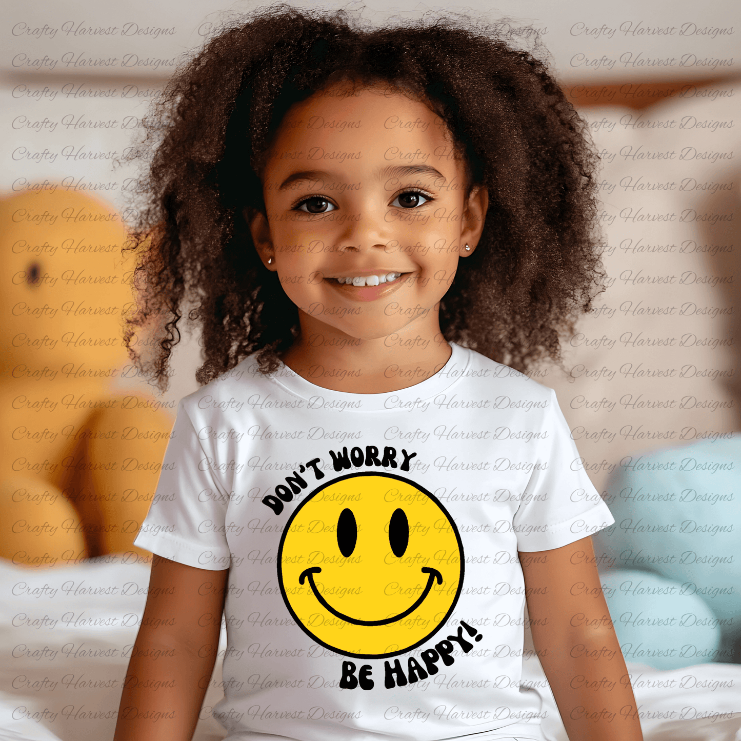 Smiling young child wearing a white t-shirt featuring a classic yellow smiley face design with the phrase ‘Don’t Worry, Be Happy!’ in black text. The background is softly blurred, showing a cozy indoor setting with plush toys and pillows.