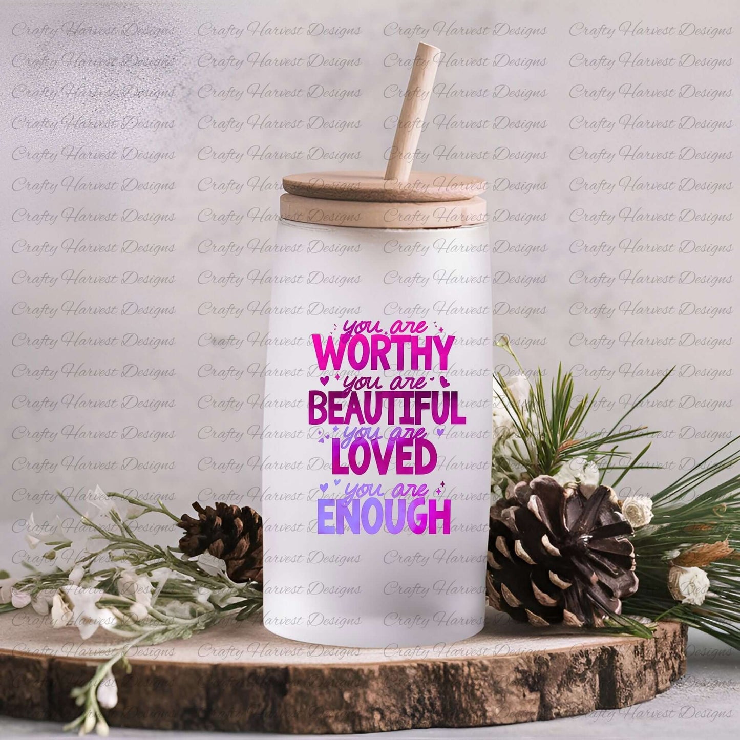 Glass can with a wooden lid and straw featuring the motivational phrase ‘You are Worthy, Beautiful, Loved, Enough’ in a gradient of pink and purple text. The glass can is displayed on a rustic surface with pinecones and greenery.