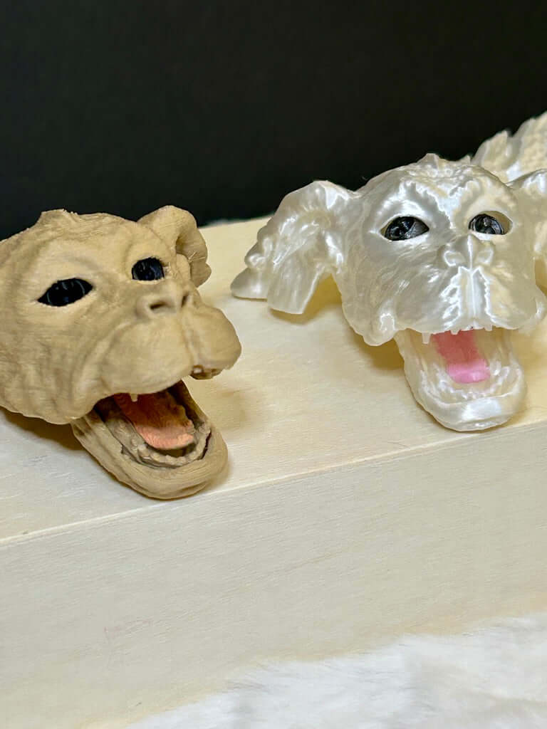 Two 3d printed dragon heads, one beige and one white with black eyes, on a light wood surface.