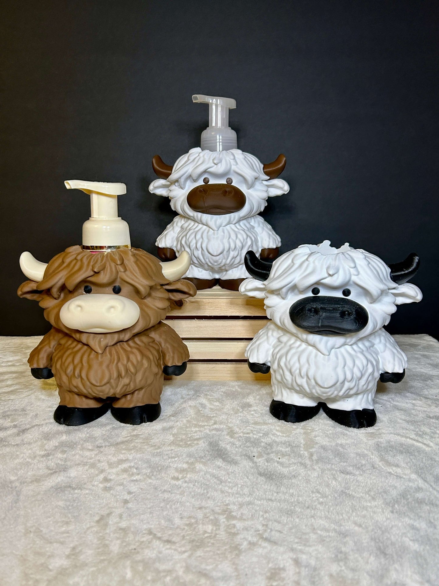 Three highland cow soap dispensers on a textured surface with a dark background
