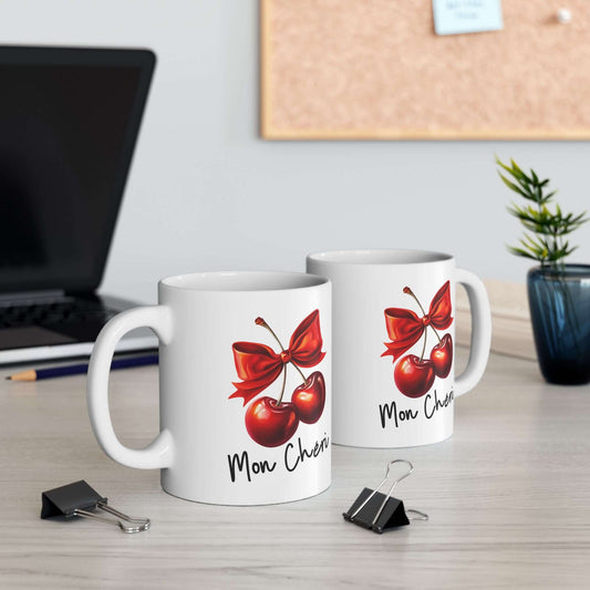 Two white ceramic mugs featuring a vibrant cherry design tied with a bold red bow and the text ‘Mon Chéri,’ displayed on a desk with office accessories.
