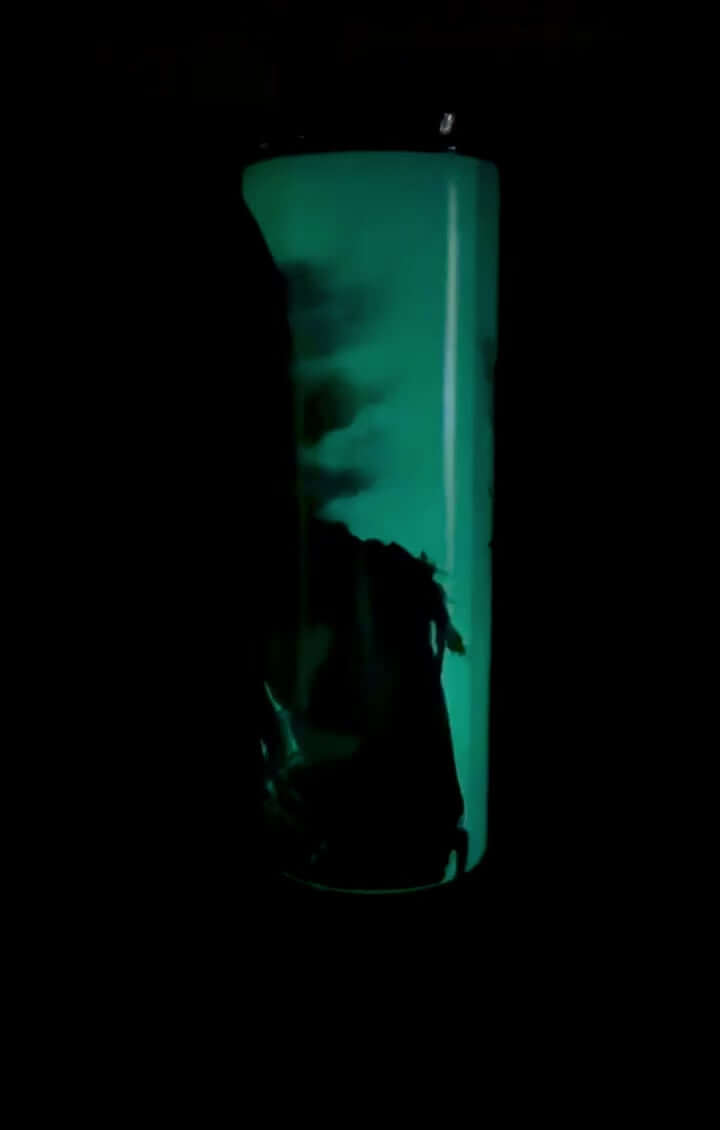 Glow-in-the-dark 20oz tumbler featuring a Grim Reaper design, illuminated in green light in a dark setting, showcasing the full wrap effect.
